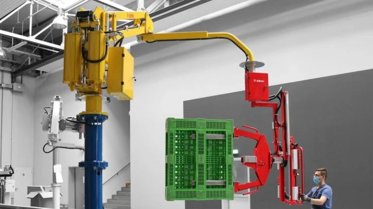 Dalmec yellow rigid-arm manipulator with red gripping tool handling a green plastic crate, operated by a worker in a packaging facility.