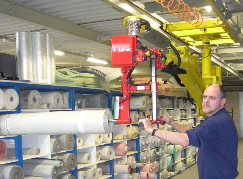 Dalmec manipulator with clamp gripping tool retrieving a fabric roll from warehouse shelving as a worker guides positioning.
