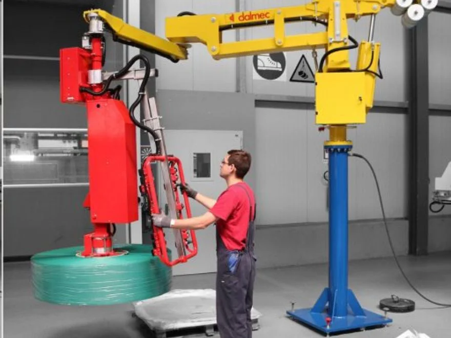 Dalmec manipulator with rotating gripping tool lifting a wrapped film reel as a worker positions the load.