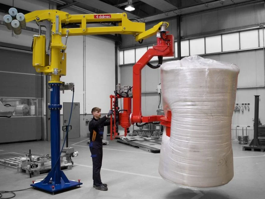 Dalmec manipulator with rotating gripping tool lifting a wrapped insulating material reel as a worker guides positioning.