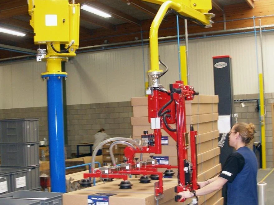 Dalmec manipulator with multi-suction cup gripping frame lifting large cardboard boxes in a warehouse, operated by a single female worker