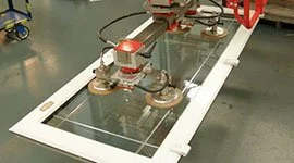 : Dalmec manipulator with vacuum gripping tool lifting a glazed window frame unit in a production facility.
