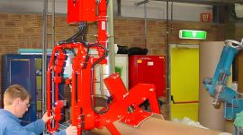 Operator using a column-mounted industrial manipulator with a custom core gripper to lift, rotate, and position a large cardboard paper tube in a manufacturing facility.