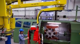Operator using a column-mounted industrial manipulator with a custom gripper to lift and position a heavy metal component at a large industrial machine in a manufacturing facility.