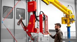 Operator using a column-mounted industrial vacuum manipulator with a multi-suction cup gripper to lift and precisely position a large glass panel in a manufacturing facility.