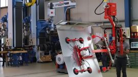 Operator using an overhead industrial vacuum manipulator with a multi-suction cup gripper to lift and position a large sheet metal panel in a manufacturing facility.